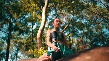 A beautiful Asian woman in a green costume sitting on a big rock while visiting the forest lot of trees inside the village video