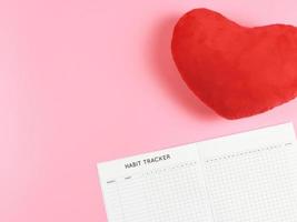 flat lay of habit tracker book with  red heart shape pillow on pink  background with copy space. photo