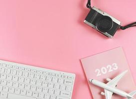 flat lay of white computer keyboard, pink diary 2023, airplane model  and camera on pink background with copy space. Travel planning concept. photo