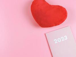 flat lay of pink 2023 diary,  red heart shape pillow on pink  background with copy space. photo