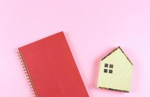 flat layout of wooden house model  and  red cover notebook  on pink  background with copy space, home purchase planning  concept. photo