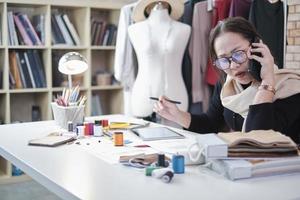 Asian middle-aged female fashion designer works in studio by talking on mobile phone about ideas and drawing sketches for dress design collection orders. Professional boutique tailor SME entrepreneur. photo