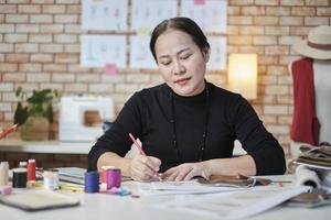 Asian middle-aged female fashion designer working in a studio by idea drawing sketches with colorful thread and sewing for a dress design collection, professional boutique tailor SME entrepreneur. photo