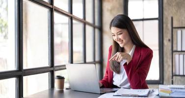 Sonríe, una mujer asiática de negocios bonita está trabajando en su trabajo en una computadora portátil en una oficina moderna. hacer un informe de análisis contable datos de inversión inmobiliaria, concepto de sistemas financieros y fiscales