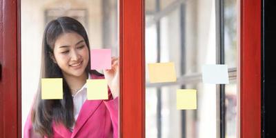 joven sonriente atractiva, mujer de negocios que usa notas adhesivas en una pared de vidrio para escribir una estrategia, un plan de negocios para el desarrollo, crecer hacia el éxito foto