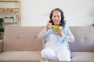 Asian senior woman sitting eating vegetable salad and healthy food and eating happily on the sofa in the house for a healthy body. healthy food concept photo