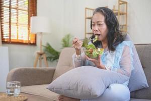 anciana asiática sentada comiendo ensalada de verduras y comida saludable y comiendo felizmente en el sofá de la casa para un cuerpo sano. concepto de comida saludable foto