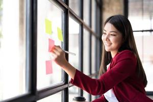 Young smiley attractive, businesswoman using sticky notes in glass wall to writing strategy business plan to development grow to success photo
