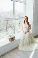 joven embarazada pelirroja con un vestido blanco cerca de la ventana foto