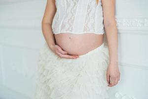 joven embarazada pelirroja con un vestido blanco cerca de la pared blanca foto