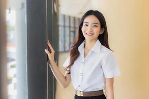 Cute Asian young woman student wear uniform is smiling and looking at camera standing to present something confidently at reading room in university background photo