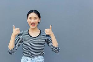 el retrato de una linda estudiante asiática tailandesa que se ve amistosa está de pie sonriendo felizmente y con confianza exitosa mostrando un golpe para la comunicación tan bueno como para presentar algo sobre un fondo gris.