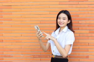 Portrait of cute Asian Thai girl student in uniform is standing smiling happily and confidently successful while holds smartphone with brick walls as background in university. photo