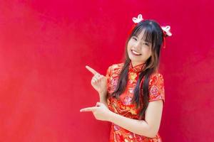 Beautiful and pretty young Asian girl with long hair who wears a red Cheongsam dress in Chinese new year theme while she shows her hand to present something on red background. photo