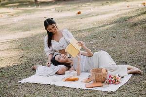 Young pretty brunette twin girls sitting on the grass with legs slightly bent in knees and looking in a brown book, wearing casual coat in autumn sunny weather on blurry background. photo