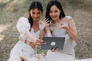 Young pretty brunette twin girls sitting on the grass with legs slightly bent in knees and looking in a brown book, wearing casual coat in autumn sunny weather on blurry background. photo