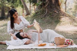 Young pretty brunette twin girls sitting on the grass with legs slightly bent in knees and looking in a brown book, wearing casual coat in autumn sunny weather on blurry background. photo