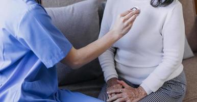 Homecare nursing service and elderly people cardiology healthcare. Close up of young hispanic female doctor nurse check mature caucasian man patient heartbeat using stethoscope during visit photo