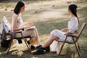 Young pretty brunette twin girls sitting on the grass with legs slightly bent in knees and looking in a brown book, wearing casual coat in autumn sunny weather on blurry background. photo
