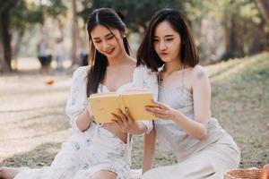 Young pretty brunette twin girls sitting on the grass with legs slightly bent in knees and looking in a brown book, wearing casual coat in autumn sunny weather on blurry background. photo