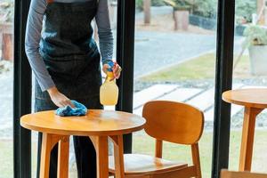 covid 19. mujer de servicio de personal de camarera asiática en mesa de limpieza de delantal con spray desinfectante para proteger la infección coronavirus en cafetería restaurante hotel. nuevo concepto normal foto