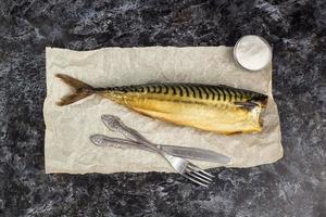 Smoked mackerel without head with fork knife on baking paper on black background photo