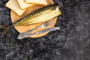 Smoked mackerel without head with fork knife cutting board bread on black background. copyspace top view photo