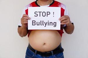 Closeup man with his big belly holds paper sign with word  STOP  Bullying . Concept, campaign for anti bullying, body shaming or parody of others physical appearance. photo