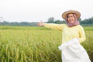 Happy Asian woman farmer is at paddy field, wear hat, yellow shirt,Thai loincloth covers  head, holds white sack of organic fertilizer, feel confident. Concept, farmer satisfied in agriculture product photo