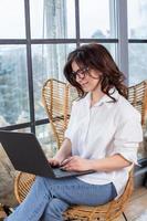 bella mujer de negocios trabajando en una laptop en una silla cerca de la ventana. mujer independiente que se conecta a internet a través de la computadora. blogger o periodista escribiendo un nuevo artículo. foto