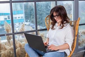 bella mujer de negocios trabajando en una laptop en una silla cerca de la ventana. mujer independiente que se conecta a internet a través de la computadora. blogger o periodista escribiendo un nuevo artículo. foto