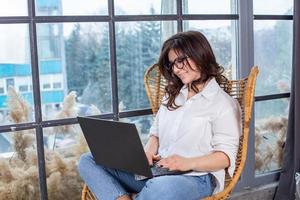 Beautiful businesswoman working on a laptop in a chair near the window. Female freelancer connecting to internet via computer. Blogger or journalist writing new article. photo