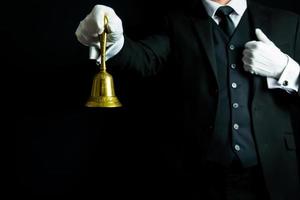 Portrait of Butler or Waiter in Black Suit and White Gloves Holding Gold Bell. Ring for Service Concept. photo