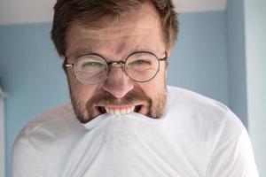 Man with glasses is angry, he tries to contain rage and bites his t-shirt. Concept of mental health. Close-up. photo