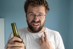 hombre desordenado señala con el dedo a su sin afeitar. el caucásico sufre de resaca, sostiene una botella de vino en las manos y hace una mueca. foto