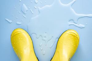 Yellow rubber boots stand near spilled water on a blue background. Concept of preparation for the rainy season. Copy space. photo