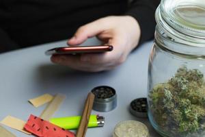 hombre sosteniendo el teléfono junto al cannabis y accesorios para fumar. comprar marihuana y hierba en línea. concepto de negocio