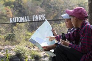 Asian young teen boys hold national park map, reading details of birdwatching before using their binoculars to watch the bird which sitting on brunches and flying in the sky, summer vacation concept. photo