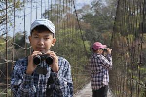 Asian boys using binoculars to watch birds on trees and fish in river in local national park during summer camp, idea for learning creatures and wildlife animals and insects outside the classroom. photo