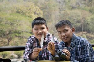 Asian boys hold bird feather and binoculars sit together in local national park to learn differentces of birds from feather during their summer vacation, leisure and outdoor activity of teens concept. photo