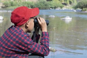 Asian boys using binoculars to watch birds on trees and fish in river in local national park during summer camp, idea for learning creatures and wildlife animals and insects outside the classroom. photo