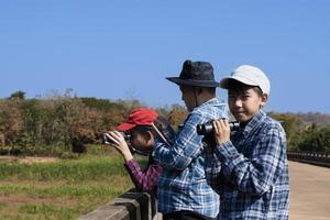 niños asiáticos que usan binoculares para observar pájaros en los árboles y peces en el río en el parque nacional local durante el campamento de verano, idea para aprender criaturas y animales salvajes e insectos fuera del aula. foto