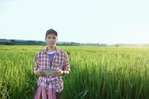 Young asian teen boy in plaid shirt, wears cap and holding tablet in hands, standing and using his tablet to survey information of rice growing and to do school project work in rice paddy field. photo