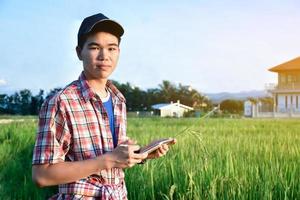 joven adolescente asiático con camisa a cuadros, usa gorra y tableta en las manos, de pie y usando su tableta para estudiar información sobre el cultivo de arroz y hacer trabajos de proyectos escolares en arrozales. foto