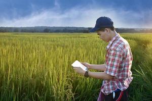 un adolescente asiático con camisa a cuadros lleva sombrero, se inclina y sostiene una espiga de arroz para comprobar la calidad del arroz y almacenar detalles en su tableta en el campo de arroz local, proyecto de arroz de los niños. foto