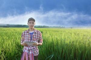 Young asian teen boy in plaid shirt, wears cap and holding tablet in hands, standing and using his tablet to survey information of rice growing and to do school project work in rice paddy field. photo