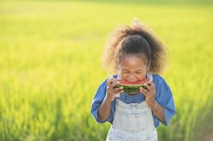 niña linda de piel negra comiendo sandía al aire libre fondo de campo de arroz verde niño africano comiendo sandía foto