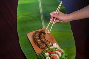 A Thai restaurant cook tastes delicious grilled herbed pork sausages from the barbeque served in the restaurant. Close-up shot of Thai sausage with fresh vegetable side dish, Thai food photo
