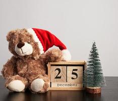 brown teddy bear in red hat, desk wooden calendar with the date december 25 and green decorative tree photo