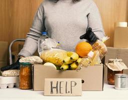 mujer con suéter gris y guantes negros sosteniendo una caja de cartón con comestibles, concepto de asistencia y voluntariado foto